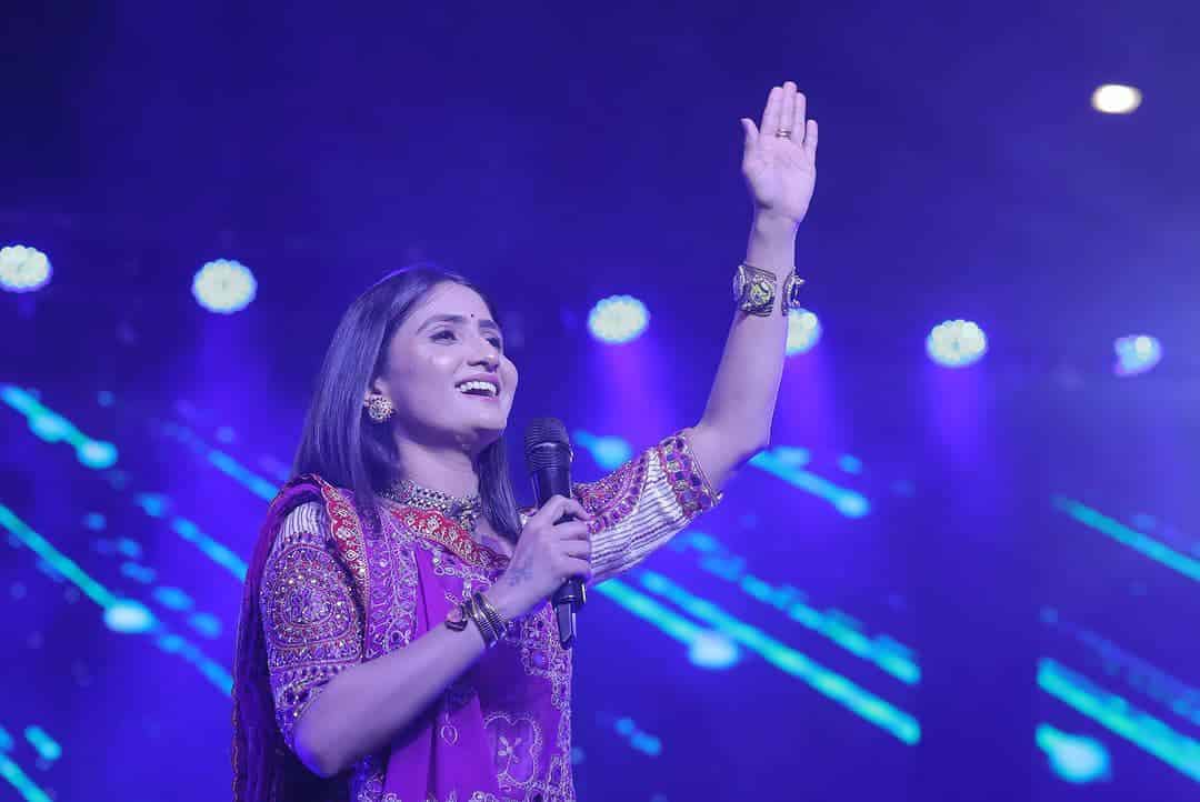 Geeta Rabari performing live in traditional Kutchi mirror-work ghagra choli during a Navratri Garba night, a large crowd dancing in traditional outfits.